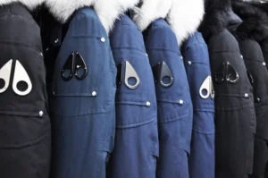 A row of winter jackets in black and blue shades, featuring fur-lined hoods and distinctive metallic teardrop-shaped logo patches on the sleeves Moose Knuckles Coats.