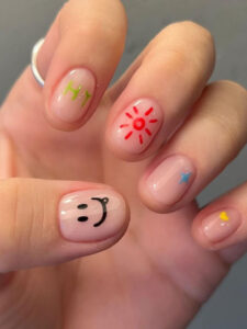 Hand with short nail designs with a clear base and colorful, minimalist designs including a smiley face, “HI” text, sun, stars, and a heart.